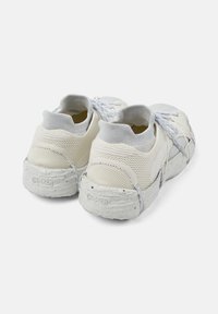 Un paio di sneakers in mesh off-white con lacci, colletto alla caviglia simile a una calza e suola texture, viste da dietro su uno sfondo semplice.