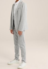 Kind, der einen hellgrauen Anzug mit Blazer und Hose, ein weißes Hemd mit Kragen und weiße Turnschuhe trägt, vor einem schlichten beigen Hintergrund steht.