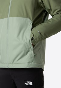 Vihreä kaksivärinen takki, jossa on sileä pinta, vetoketju edessä, taskut ja "DRYVENT"-merkki. Yhdistetty mustiin housuihin.