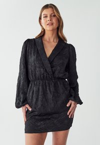Schwarzes Kleid mit strukturiertem, zerknittertem Stoff, tiefem V-Ausschnitt, puffartigen langen Ärmeln und einem elastischen Taillenbund für eine figurbetonte Silhouette.