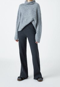 Pull en maille grise avec un col montant, assorti à un pantalon évasé gris foncé et des chaussures noires à enfiler avec des accents pailletés. Tissu texturé.