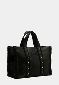 Sac fourre-tout noir avec des côtés en fausse fourrure, un centre en cuir lisse, un logo embossé et des clous argentés pour les détails. Poignées doubles pour le transport.