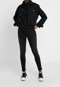 Giacca in denim nera con chiusure a bottone e tasche, dolcevita nero, jeans skinny neri e sneakers nere con suola bianca.