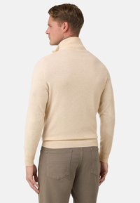 Pull en tricot beige avec un col montant, des bords côtelés et une texture douce, associé à un pantalon marron clair avec des poches arrière.