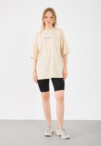 Beige oversized t-shirt met korte mouwen en een zwart gedrukt ontwerp, gecombineerd met zwarte fietsshorts en lichtgekleurde sportschoenen.