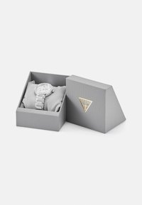 Sølvklokke med rund urskive, metalllenke armbånd og dekorative detaljer i en grå boks med en trekantet Guess-logo på forsiden.