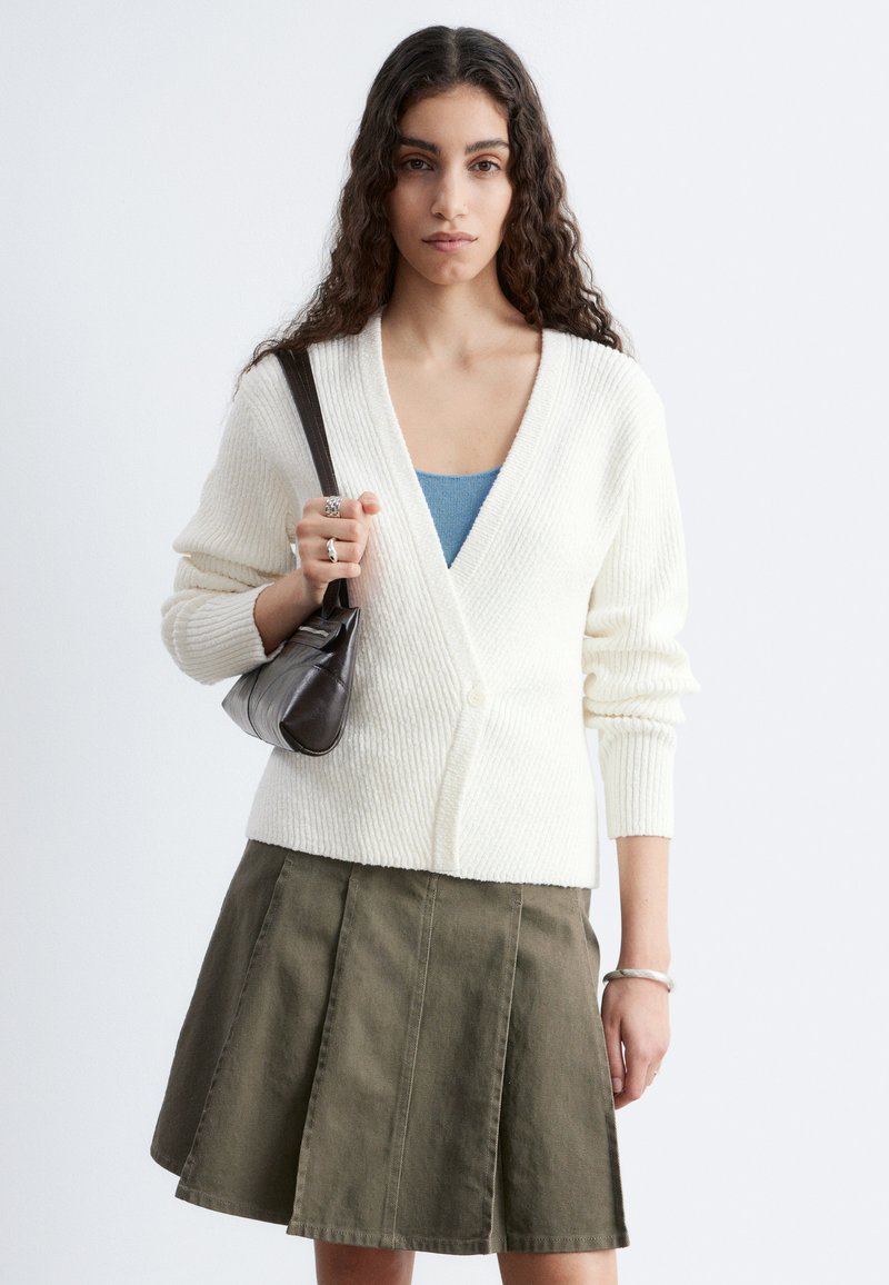 Femme aux longs cheveux bouclés portant un cardigan en maille blanc, un haut bleu, une jupe plissée olive, tenant un sac à épaule noir devant un fond uni.
