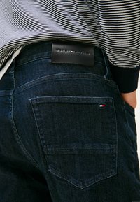 Donkerblauwe denim jeans met een strak ontwerp, voorzien van een leren label met de opdruk "Tommy Hilfiger" en een klein merkembleem op de achterzak.