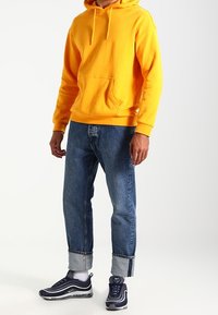 Uomo che indossa un felpa gialla brillante, jeans blu risvoltati, calzini bianchi e sneakers neri e bianchi Nike mentre sta in piedi su uno sfondo bianco.