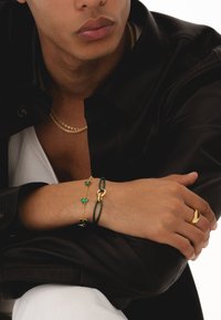 Bracelet en chaîne dorée avec accents verts, bracelet en cuir avec quincaillerie dorée, anneau doré, colliers en or superposés, veste foncée, chemise claire.
