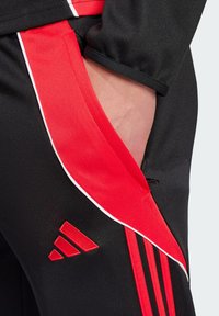 Zwarte sportbroek met rode zijpanelen en witte piping. Beschikt over een geborduurd rood logo en een zak aan de zijkant.