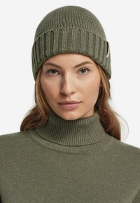 Olivgrön stickad mössa med ribbat mönster, försedda med en liten accentetikett. Matchad med en olivgrön ribbad polotröja.