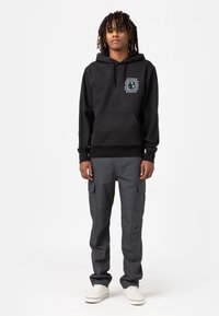 Zwarte hoodie met voorzak, met een witte grafische logo. Gedragen met grijze cargo-broeken, die meerdere zakken tonen, en witte sneakers.