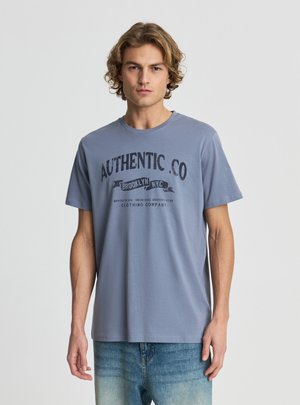 Młody mężczyzna z kręconymi włosami, ubrany w niebieską koszulkę z napisem „AUTHENTIC .CO” oraz wyblakłe niebieskie dżinsy, stojący na tle jednolitego jasnego tła.