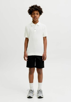 Băiat cu păr creț stă îmbrăcat într-un tricou polo alb cu mâneci scurte, pantaloni scurți negri, șosete albe și adidași gri pe un fundal alb.