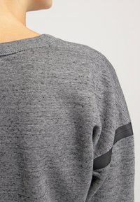 Grå tröja med texturerad tyg, ribbad halsringning och en svart rand som accent på ärmen. Nära bild av bakvyn.