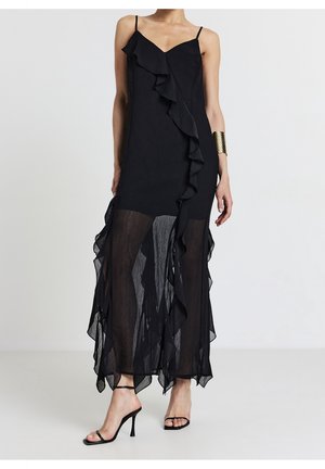 Kobieta w długiej czarnej sukience bez rękawów z falbanami i przezroczystą tkaniną od połowy uda w dół, w połączeniu z czarnymi sandałami na wysokim obcasie i złotą bransoletą na nadgarstku.