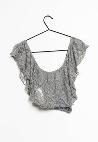 Grå spetsblus med breda fyrkantiga ärmar och rundad halsringning, som visar upp blommönster och en något genomskinlig textur. Hänger upphängd på en svart krok.