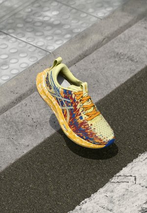 Chaussure de running jaune et orange avec des accents bleus reposant sur le trottoir et le pavage texturé près d'un passage pour piétons.