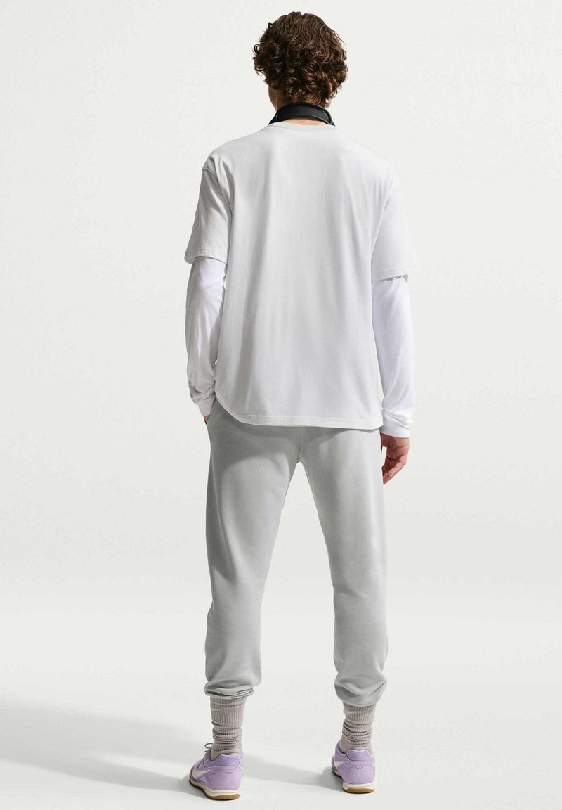 Hombre con pelo rizado de espaldas, lleva puestas camisetas blancas superpuestas de manga larga y corta, pantalones deportivos grises, calcetines grises y zapatillas moradas.