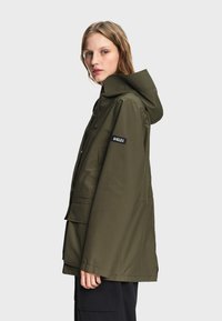 Jeune femme portant une veste de pluie à capuche vert olive avec un patch logo "Aigle" sur la manche, légèrement de profil sur un fond uni.