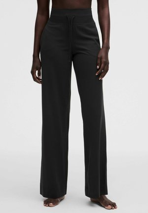 Persona che indossa pantaloni neri a vita alta con gamba ampia e coulisse, in piedi a piedi nudi su uno sfondo semplice e chiaro.
