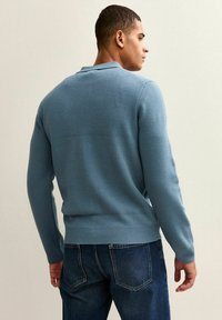 Ljusblå stickad tröja med rund krage, ribbade ärmslut och midja. Buren med mörka denimjeans, i en lätt vinkel från baksidan.
