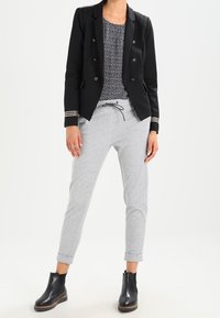 Černý přiléhavý blazer se zlatými knoflíky v detailech, kombinovaný se šedými jogger kalhotami. Na sobě má námořnickou modrou blůzu s bílými puntíky a černé kotníkové boty.