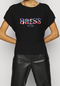 Schwarzer Strickpullover mit kurzen Ärmeln, verziert mit bunten, paillettenbesetzten "GUESS"-Schriftzügen und "EST. 1981" in Blau, kombiniert mit schwarzen Lederhosen.