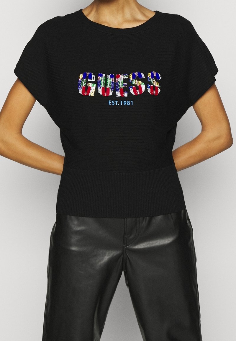 Schwarzer Strickpullover mit kurzen Ärmeln, verziert mit bunten, paillettenbesetzten "GUESS"-Schriftzügen und "EST. 1981" in Blau, kombiniert mit schwarzen Lederhosen.
