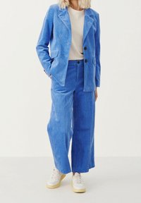Blazer blu in corduroy e pantaloni a gamba larga abbinati a un top beige e sneaker bianche. Il tessuto ha un aspetto testurizzato e a coste.