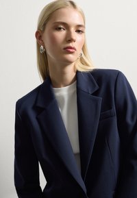 Blazer navy con rever strutturati e taschino sul petto, abbinato a un top bianco strutturato. La modella indossa orecchini metallici.