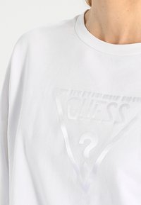 Sudadera de algodón blanca con un logo triangular brillante que incluye el texto "GUESS" y un signo de interrogación, cuello redondo y mangas cortas.