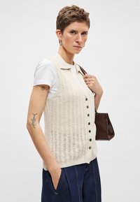 Πλεκτό γιλεκό σε μπεζ χρώμα, με γιακά, κουμπιά στο μπροστινό μέρος και υφασμένο μοτίβο. Συνδυασμένο με λευκό T-shirt και σκούρο τζιν παντελόνι.