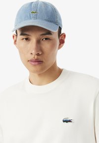 Cappello in denim di colore azzurro chiaro con logo di coccodrillo ricamato in verde, abbinato a una t-shirt in cotone bianco con un piccolo logo sul petto.