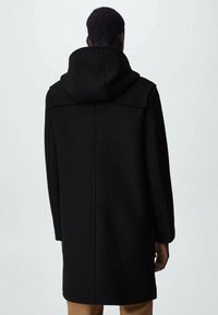 Manteau en laine noire avec capuche, présentant une coupe droite, des détails cousus et des manches longues, s'étendant juste au-dessus des genoux.