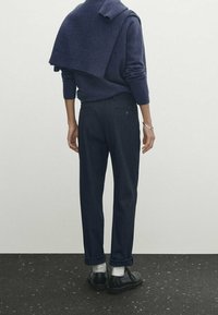 Marinblå ulltröja draperad över axlarna, i kombination med mörka byxor. Har rullade ärmslut och svarta skor med vita strumpor. Enkla, rena linjer.