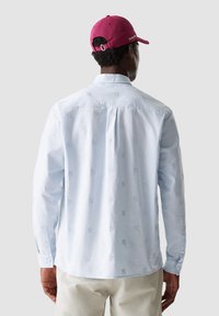 Camicia azzurro chiaro a maniche lunghe con motivo logo ricamato, polsini con bottone e dettaglio sul retro. Indossata con un cappellino bordeaux.