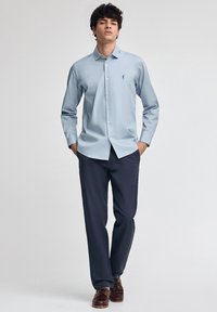 Giovane in piedi con le mani in tasca, che indossa una camicia azzurro chiaro, pantaloni blu scuro e scarpe di pelle marroni su uno sfondo neutro.
