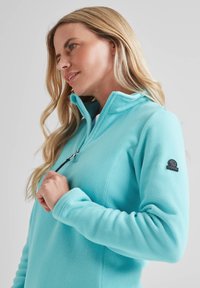 Turquoise fleece pullover with a high collar and half-zip closure. It features a smooth texture and a logo accent on the sleeve.