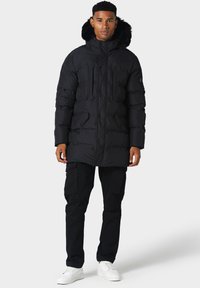 Zwarte donsjack met een capuchon gevoerd met bont, een ritssluiting aan de voorkant en meerdere zakken aan de voorkant. Gedragen over cargobroek en witte sneakers.