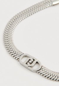 Κοντινό πλάνο από ένα ασημένιο κολιέ με αλυσίδα φίδι και οβάλ κούμπωμα που διαθέτει σχέδιο με αλληλοσύνδετους στρόβιλους.