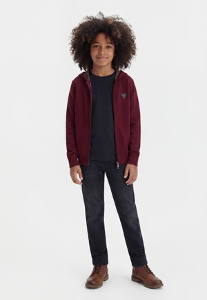 Enfant aux cheveux bouclés portant un sweat à capuche bordeaux zippé, un t-shirt noir, un jean foncé et des bottes marron, debout sur un fond blanc.