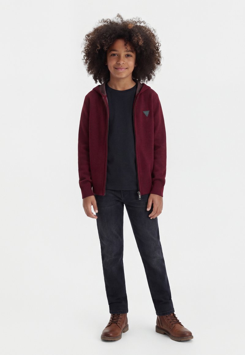 Enfant aux cheveux bouclés portant un sweat à capuche bordeaux zippé, un t-shirt noir, un jean foncé et des bottes marron, debout sur un fond blanc.