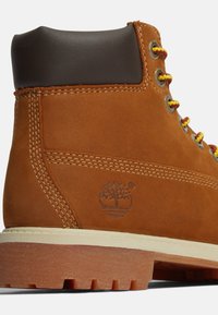 Stivale in suede marrone con un colletto in pelle marrone scuro, lacci gialli e rossi, e una suola in gomma testurizzata con un logo ad albero.
