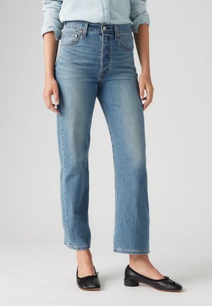 Femme portant un jean droit taille haute bleu clair, une chemise à manches longues bleu clair et des ballerines noires à talons bas, debout sur fond blanc.