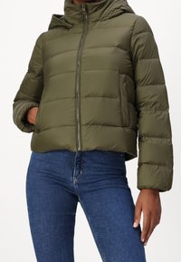 Giacca imbottita verde oliva con cappuccio, caratterizzata da una chiusura frontale con zip, design trapuntato e polsini elasticizzati. Abbinata a jeans blu.