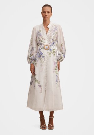 Kvinne iført en hvit midi-kjole med blomsterprint, puffede lange ermer, knappelukking foran og belt med rund spenne, kombinert med hvite hæler.