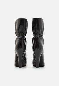 Μαύρες ψηλές μπότες μέχρι τον αστράγαλο από λείο δέρμα, με πτυχώσεις στην πάνω πλευρά, στρογγυλή μύτη και κομψό σχέδιο τακουνιού, που αναδεικνύουν μια μινιμαλιστική αισθητική.