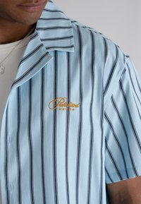 Light blue short-sleeve shirt with black vertical stripes, featuring an embroidered "Prohibited" logo in gold on the chest.